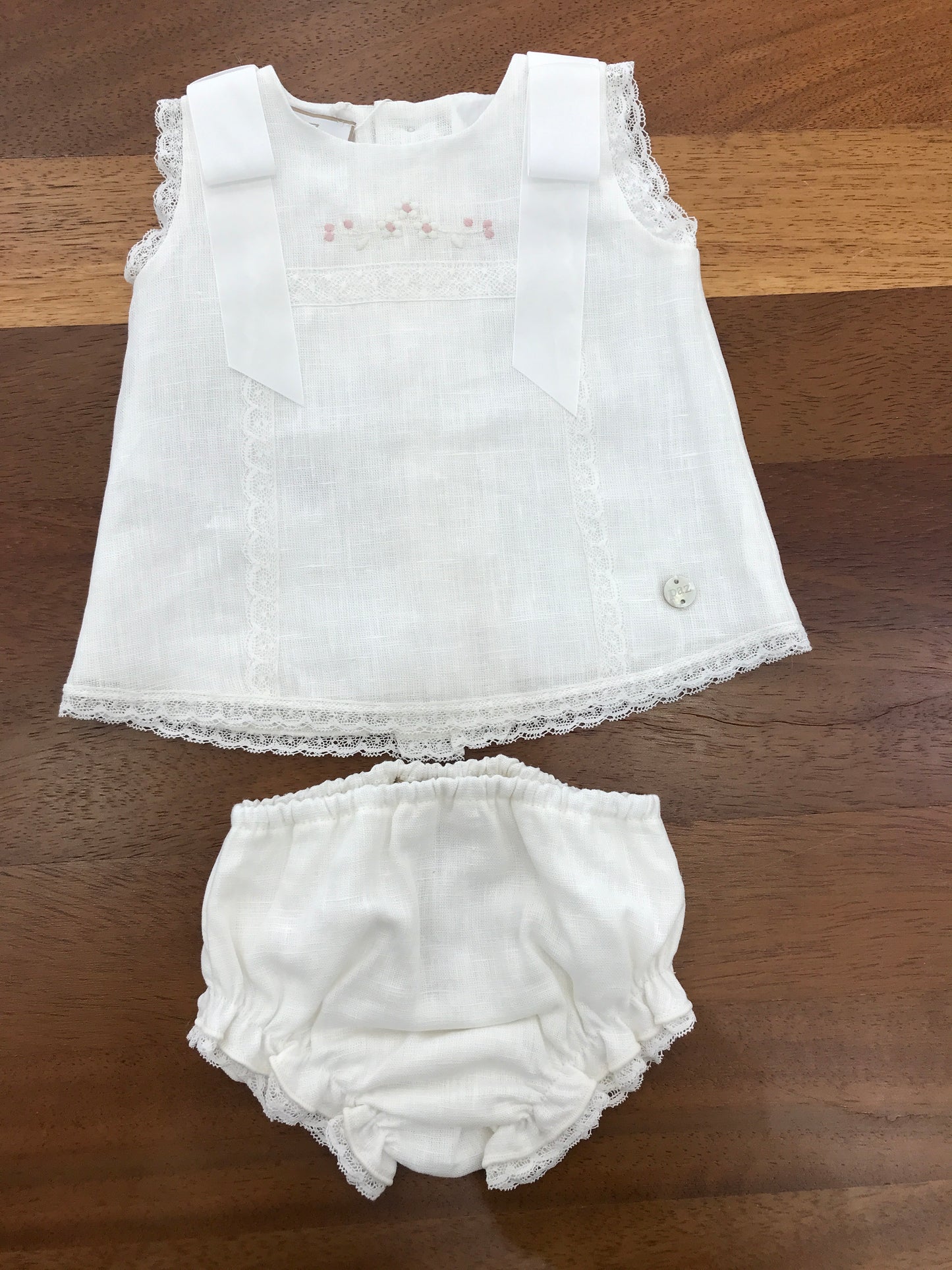 Completo Paz Rodriguez in lino bianco con fiocchi sulle spalle con merletto con fiori ricamati sul davanti con culotte e cuffia abbinata