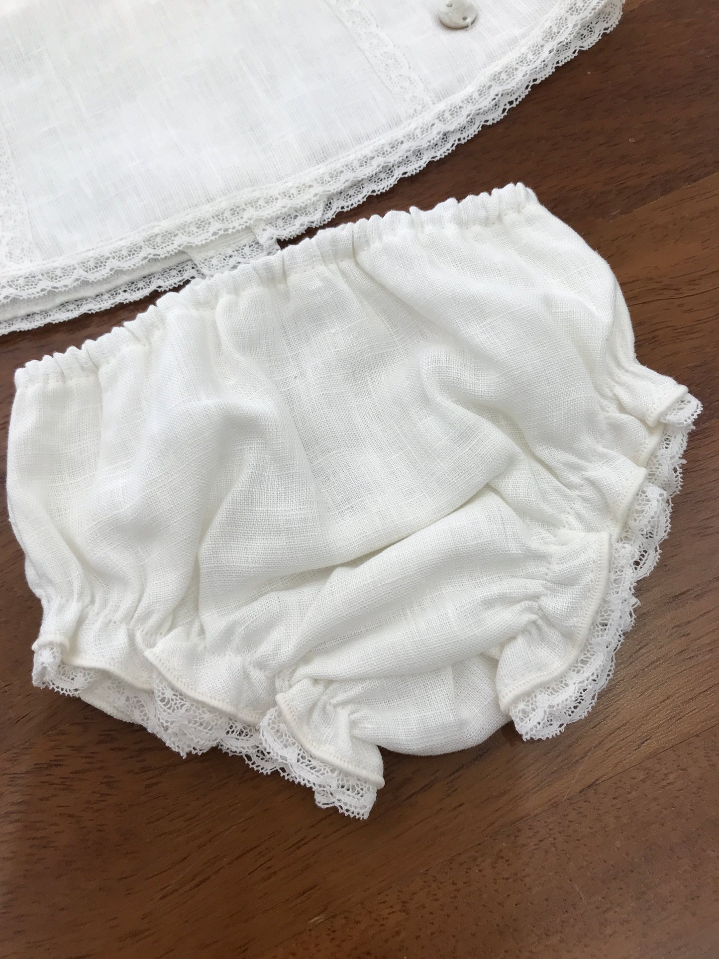 Completo Paz Rodriguez in lino bianco con fiocchi sulle spalle con merletto con fiori ricamati sul davanti con culotte e cuffia abbinata